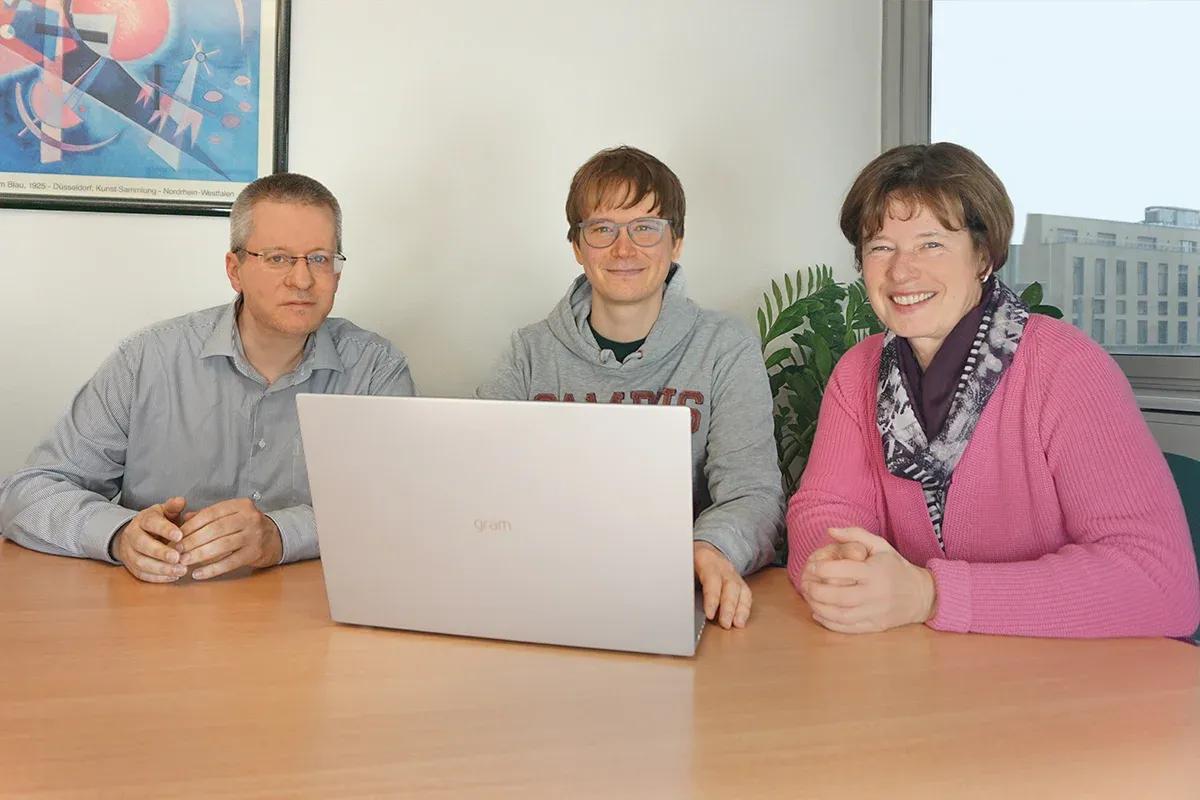 Three people, two adults and one younger, collaborate around a laptop in an office.